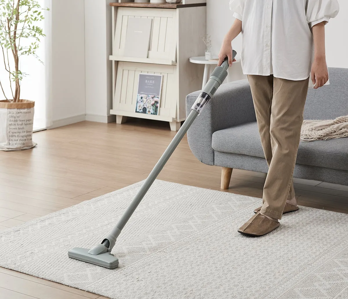 掃除機をかけている女性の画像。女性は白いシャツと茶色のズボンを着用し、灰色のカーペットの上で掃除機をかけている。背景には、本棚、ソファ、観葉植物がある。