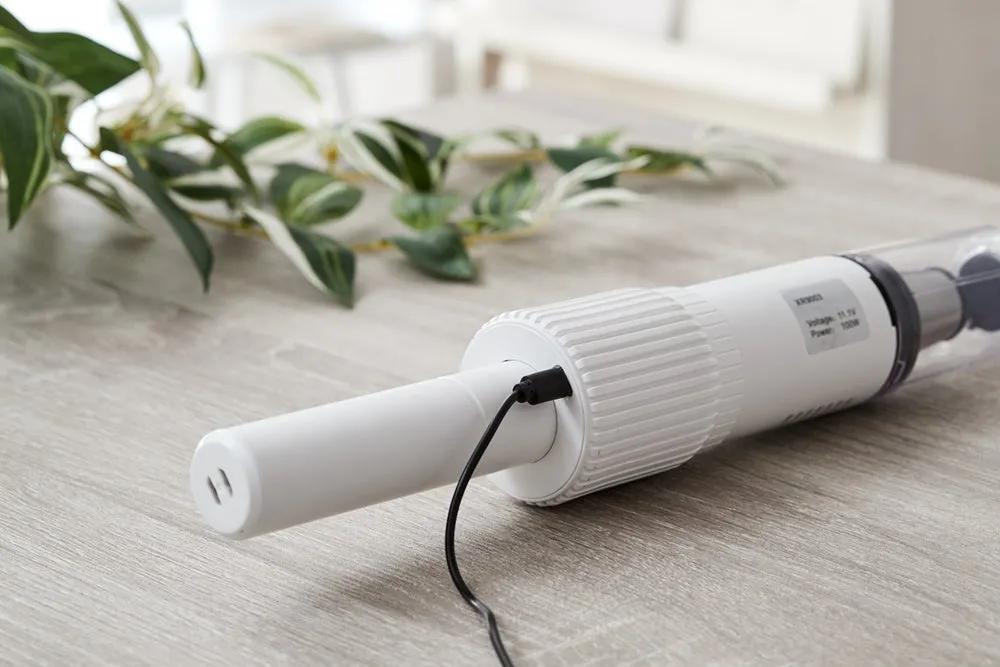 木製のテーブルの上に置かれた白いコードレス掃除機のクローズアップ写真。掃除機は充電中であり、テーブルには葉の装飾が施されている。