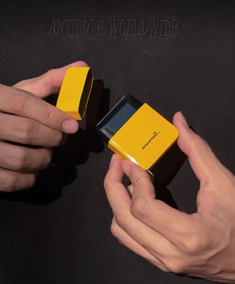 2つの手に持たれた黄色い箱のクローズアップ写真。片方の手は箱の蓋を持ち、もう片方の手は箱の本体を持っています。背景は黒い布です。