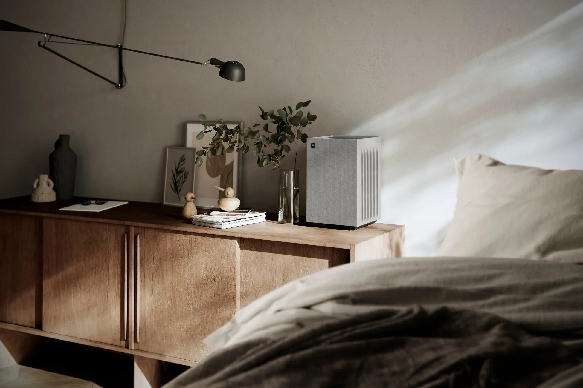 木製のサイドボードの上に置かれた空気清浄機と装飾品が写った写真。ベッドが手前にあり、壁には照明がある。