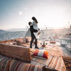 男性が女性を抱き上げている様子。背景には街並みと夕焼けが見える。