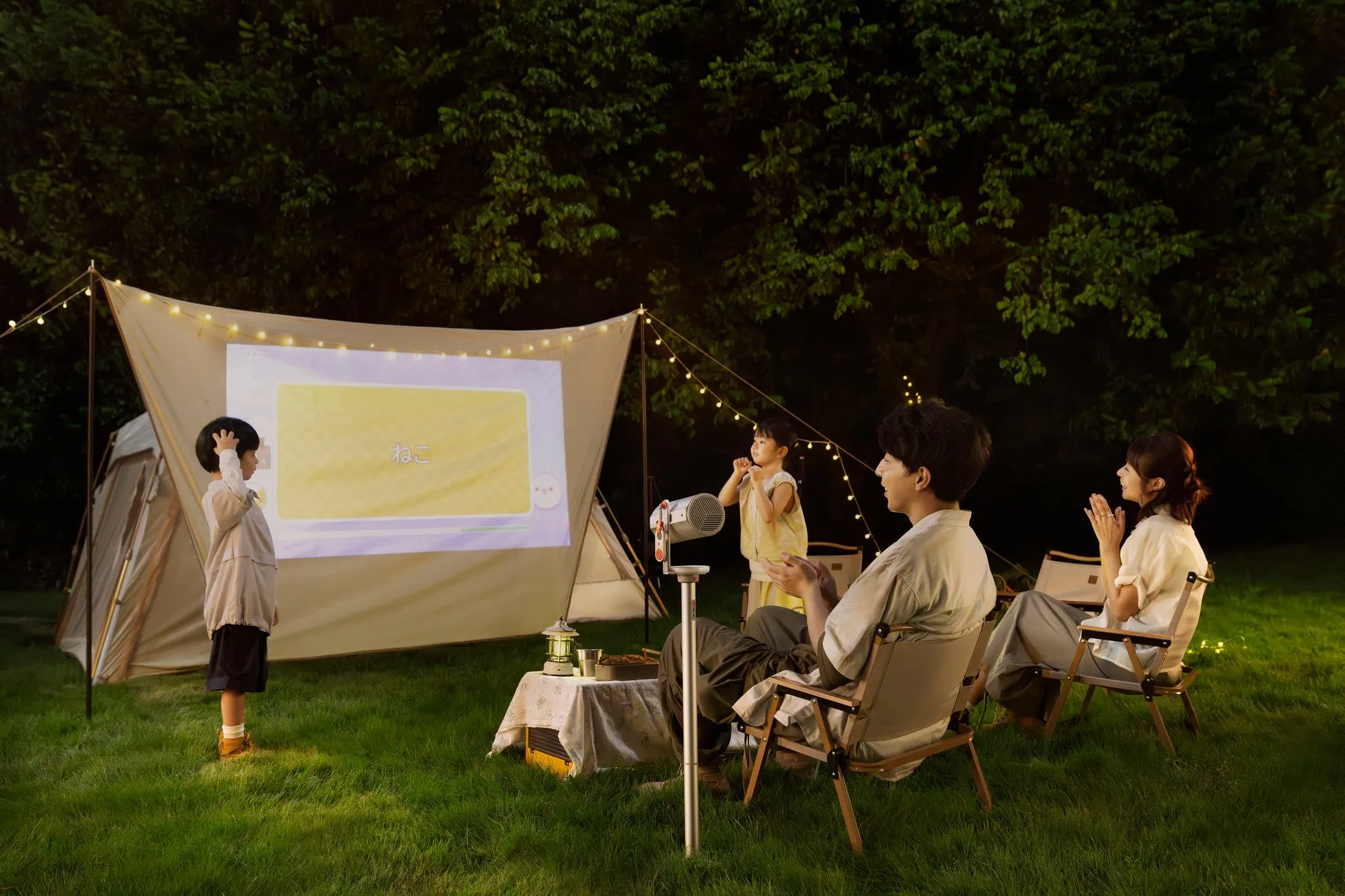 庭で家族がプロジェクターを使って映画を見ている様子。子供たちはスクリーンを見ており、両親は椅子に座って楽しんでいる。