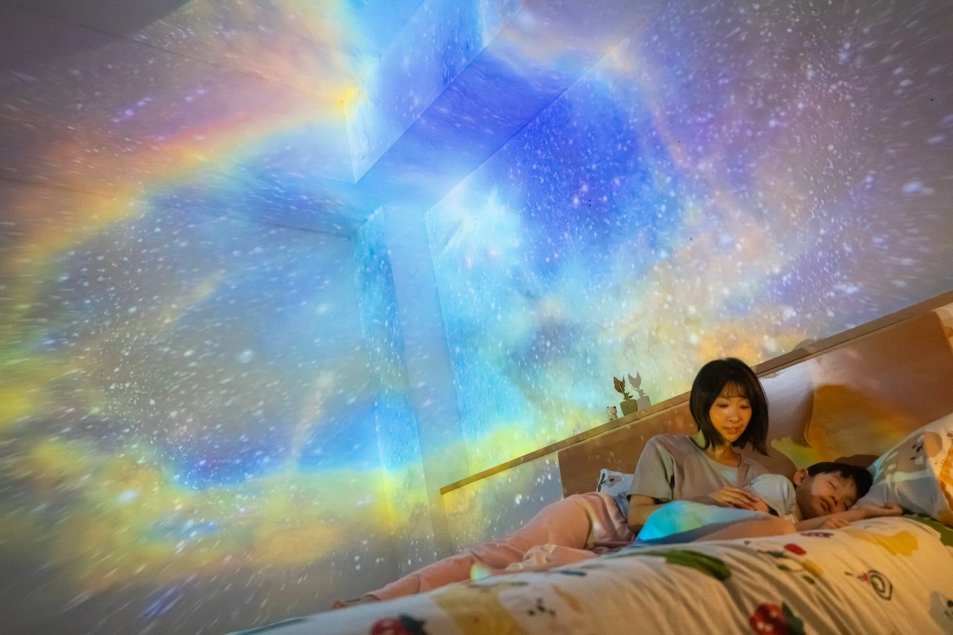 星空が投影された部屋で、母親と子供が一緒にベッドで眠っている様子を捉えた写真です。天井と壁には星空が映し出され、幻想的な雰囲気を醸し出しています。母親は子供に寄り添い、優しく見守る姿が印象的です。
