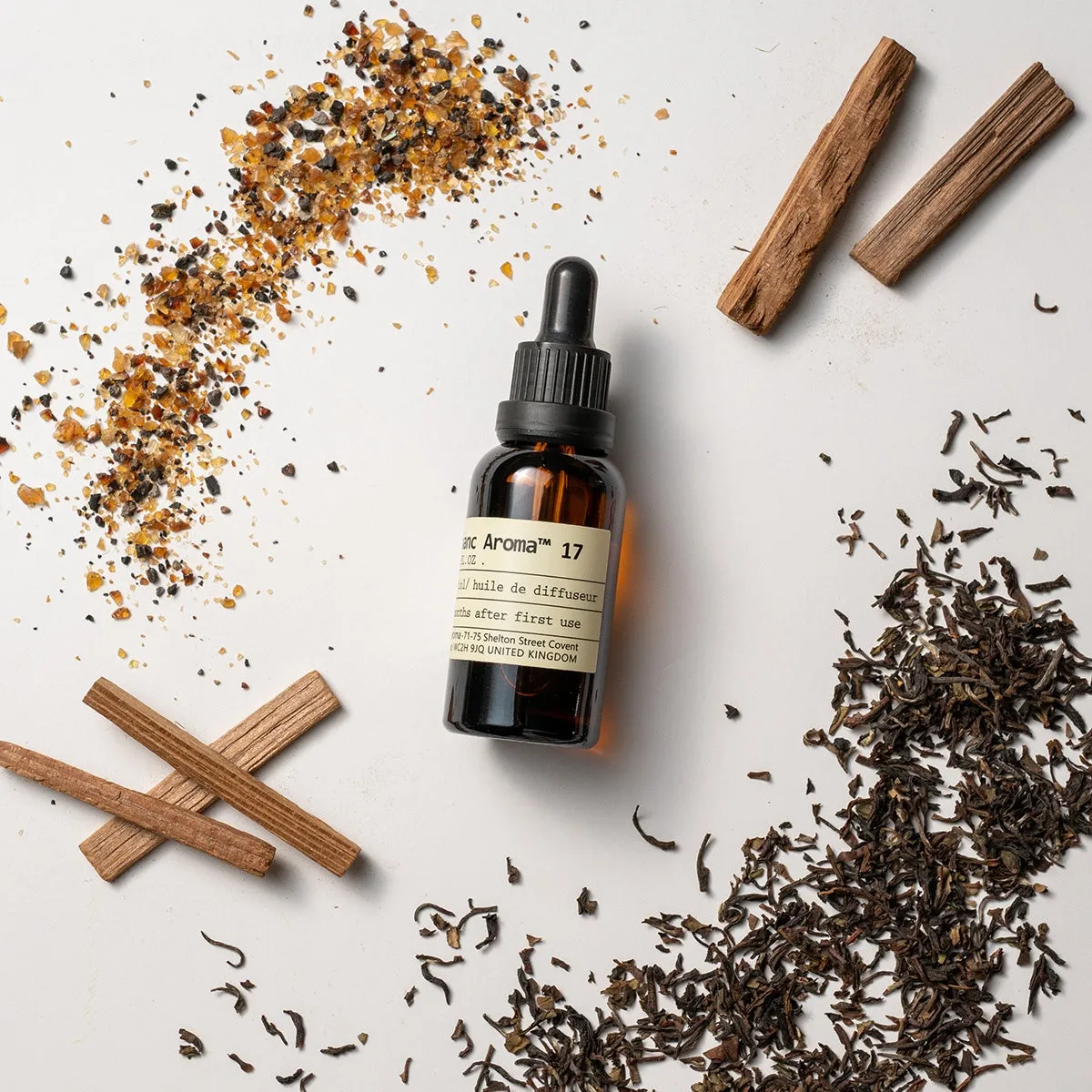 茶色のガラス瓶と、木片、茶葉、香料が散らばっている様子を真上から捉えた写真です。瓶には黒いキャップとラベルが貼られています。背景は白です。