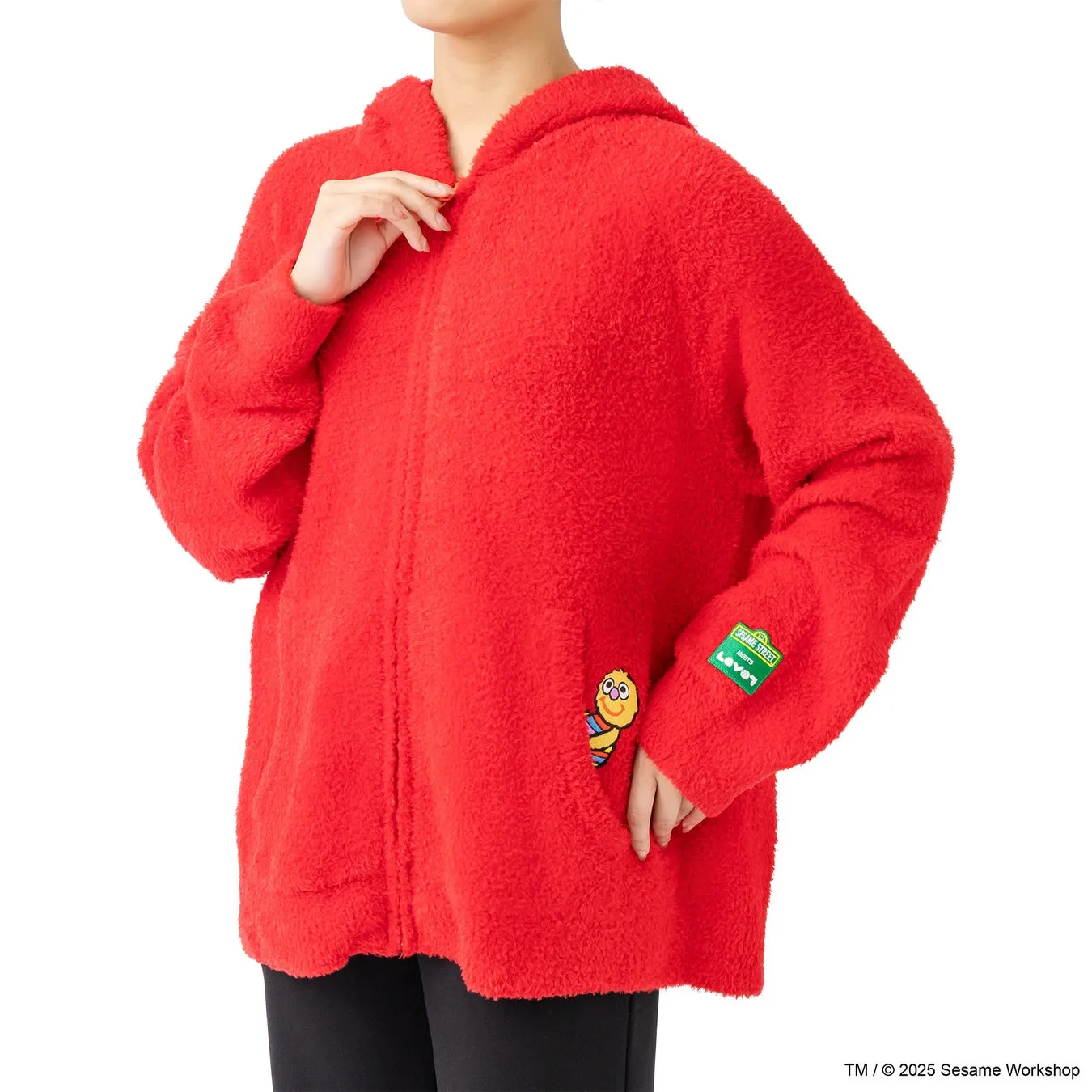 赤いフリースパーカーを着た女性の写真。パーカーにはセサミストリートとレベッツのロゴが付いています。女性は黒いズボンを履いています。