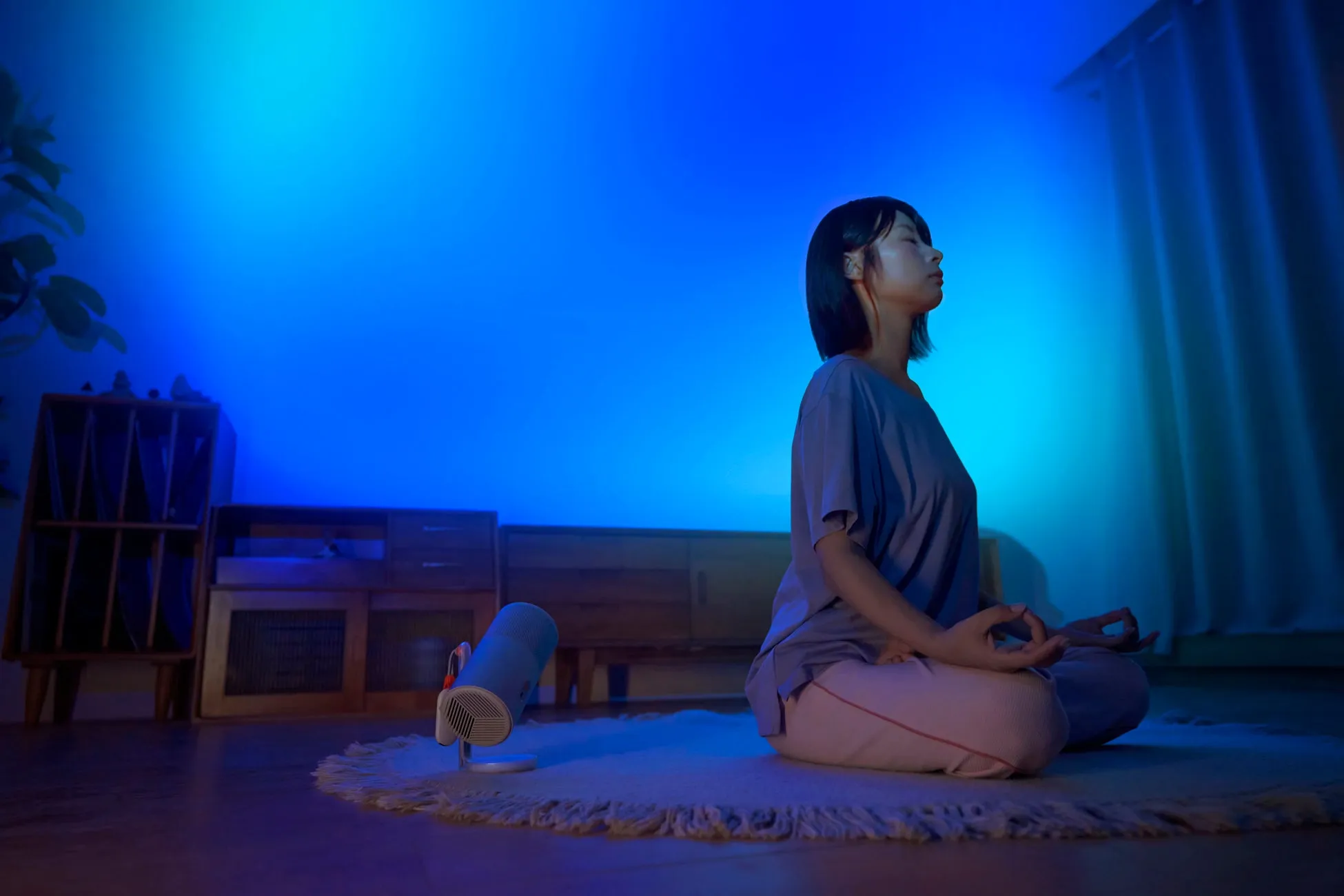 女性が瞑想している写真。青い光に照らされた部屋で、床に座り、目を閉じています。部屋には、植物、収納棚、加湿器のようなものが写っています。