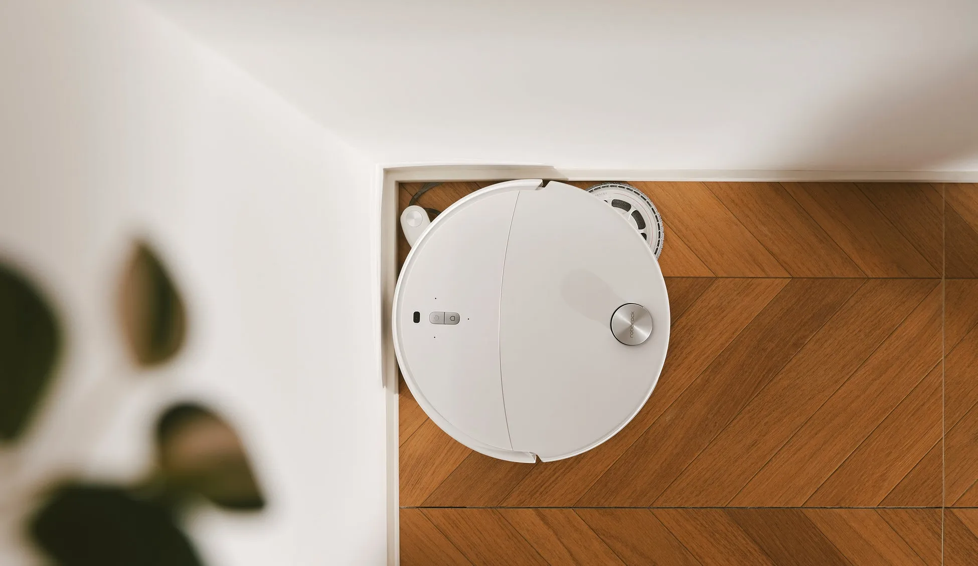 白いロボット掃除機が木製の床に置かれています。上部からのショットで、背景に白い壁があります。