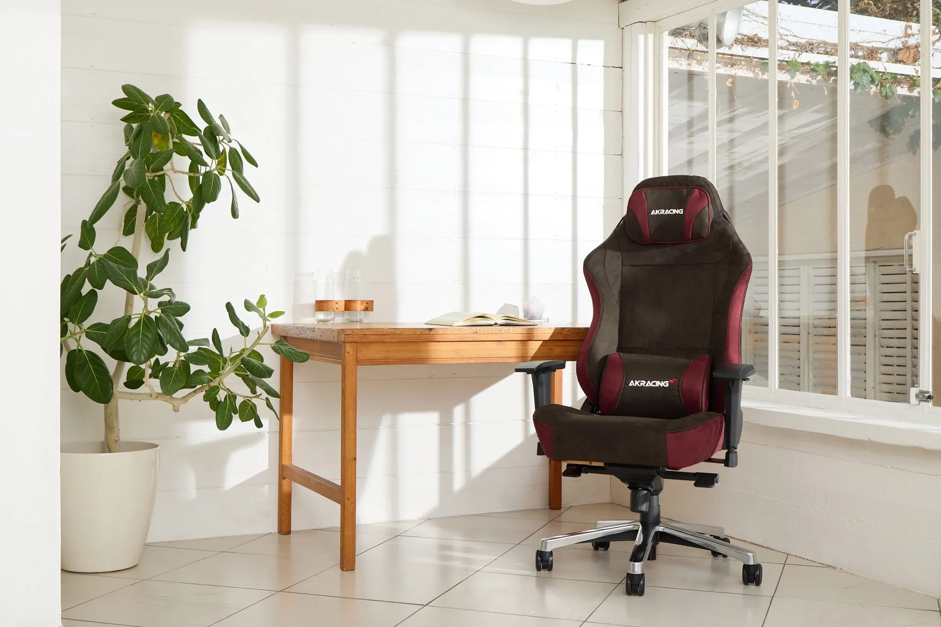 茶色と赤のゲーミングチェアが置かれた、明るい窓辺の部屋の写真です。テーブルには本とグラスがあり、観葉植物も配置されています。
