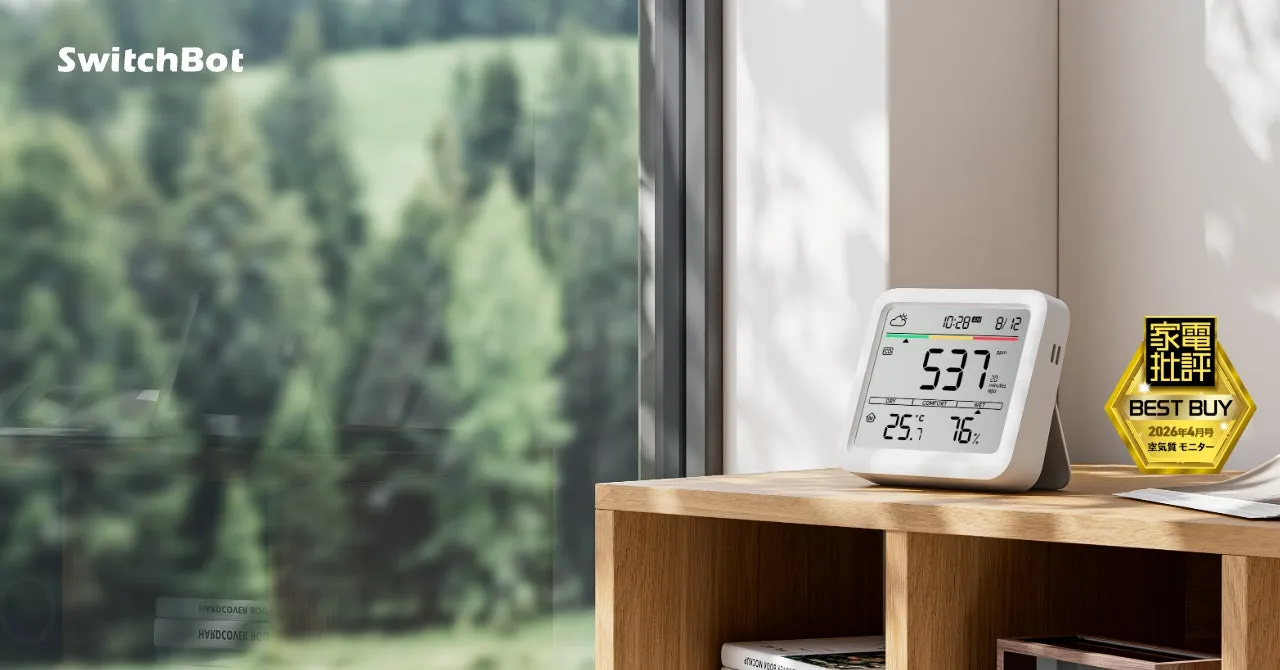 SwitchBotの空気質モニターが置かれた、木製の棚と窓辺の風景の写真。モニターには時刻、日付、空気質指数、温度、湿度が表示されている。BEST BUYのステッカーが貼られている。