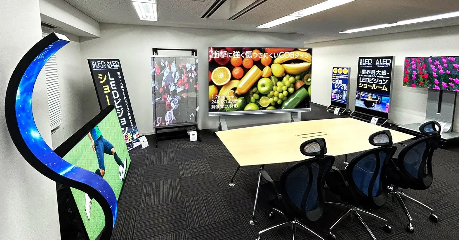 LEDビジョンが展示されたショールームの様子。会議室のような空間に、様々なサイズのLEDディスプレイが配置され、映像が映し出されている。中央には会議用のテーブルと椅子が並び、LEDビジョンに関する情報を表示した看板も見える。