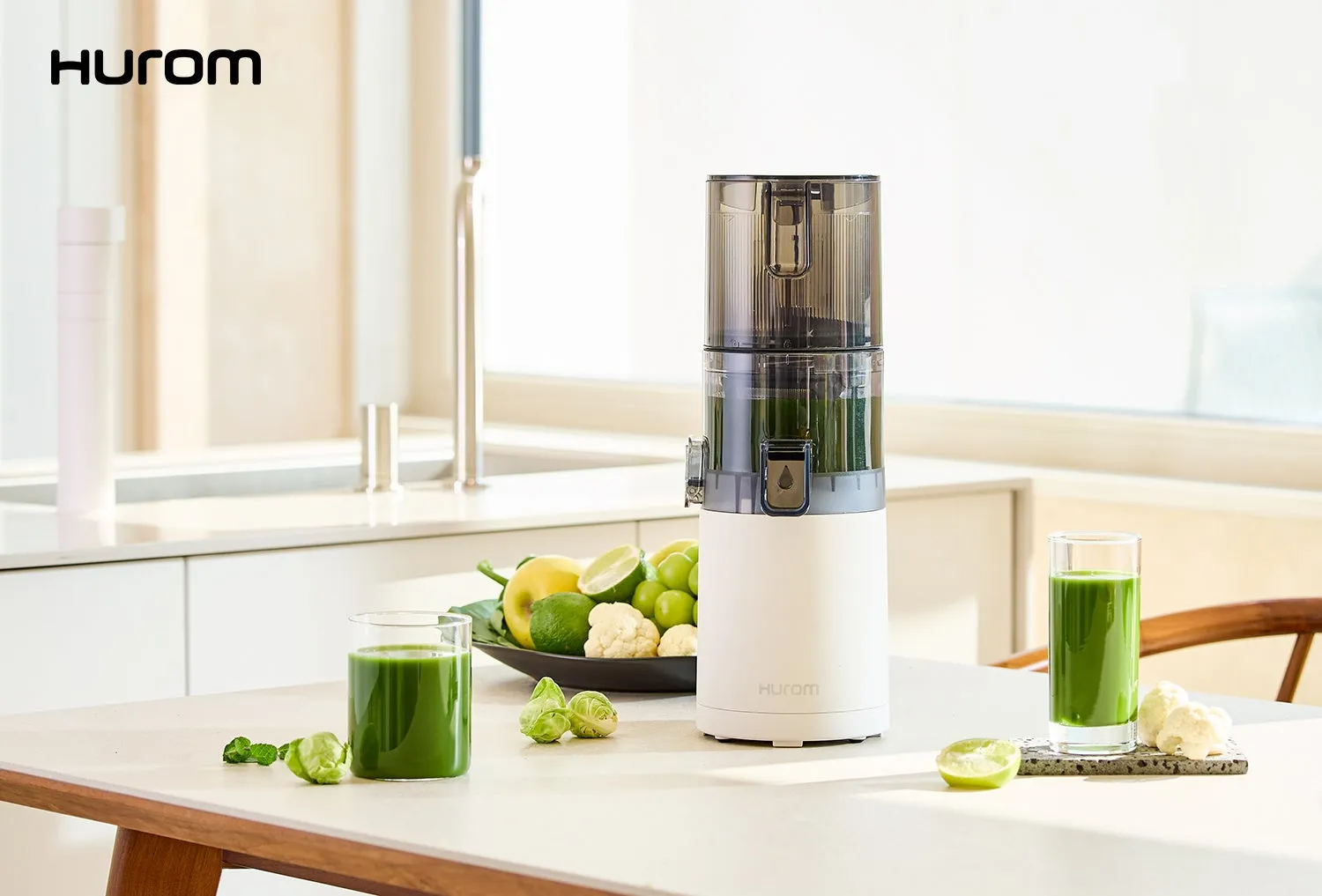 ジューサーとグラスに入ったグリーンスムージーがテーブルの上に置かれています。テーブルには、カットされたライム、ブロッコリー、カリフラワー、リンゴなどの果物と野菜の盛り合わせもあります。