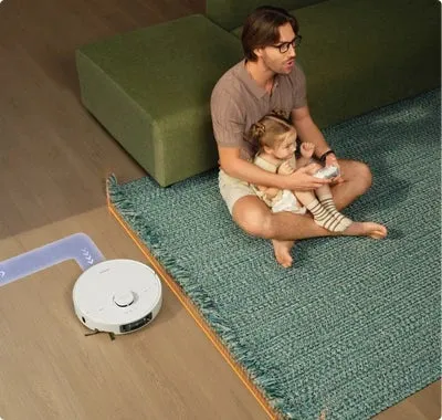 父親が娘を膝に乗せて座り、ロボット掃除機が床を掃除している様子