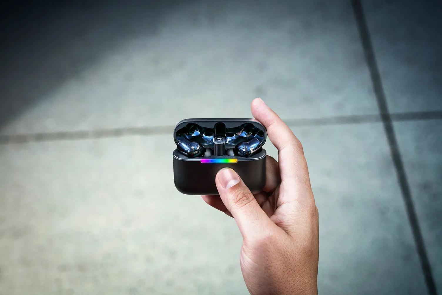 黒色のイヤホンが入ったケースを手に持っている様子を捉えた写真です。ケースには虹色のライトが点灯しています。背景はグレーのコンクリートです。