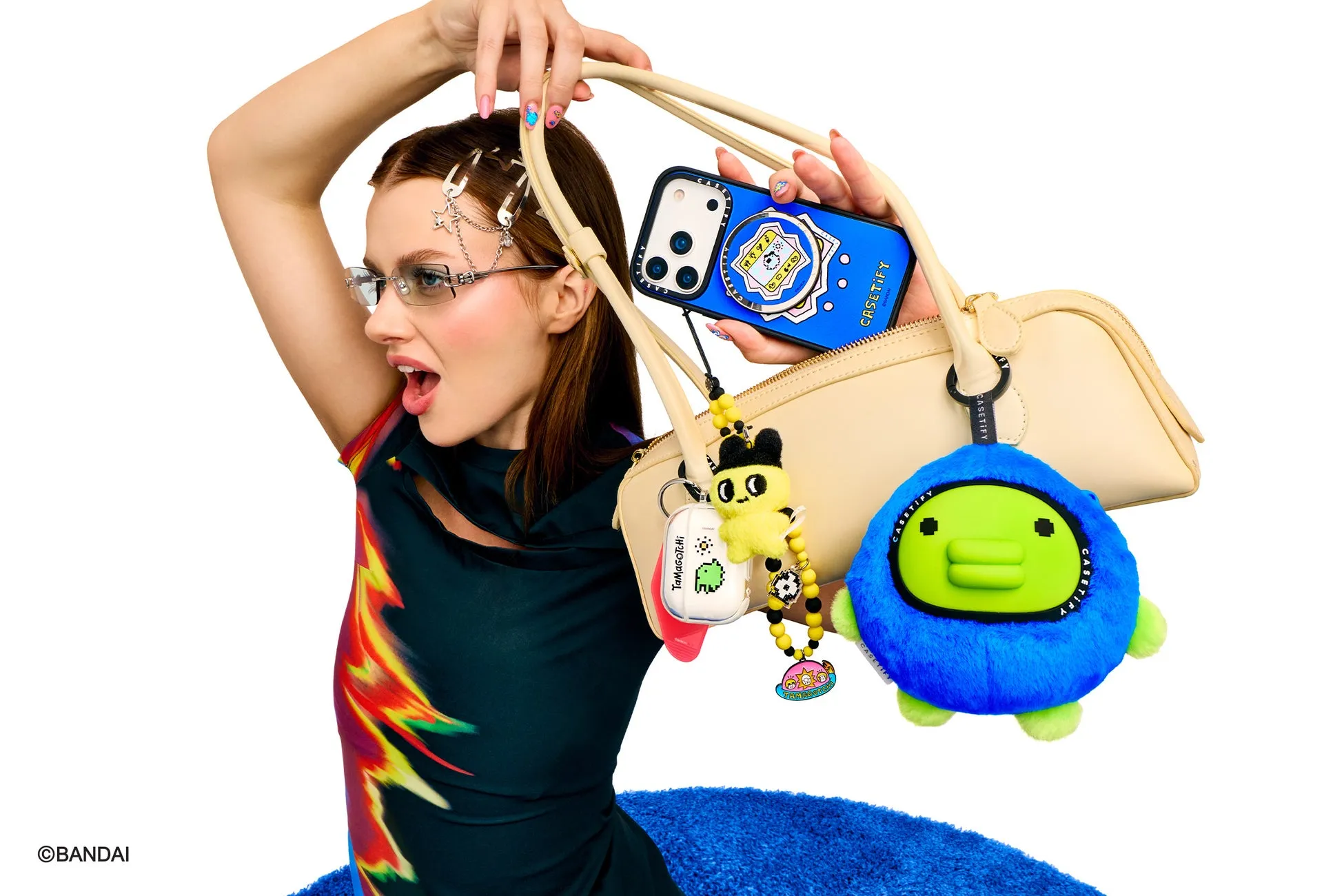 女性がバッグを持ち、スマートフォンケースやアクセサリーを見せている写真。彼女はメガネをかけ、驚いたような表情をしている。バッグにはTamagotchiのアクセサリーやCASETiFYのケースが付いている。